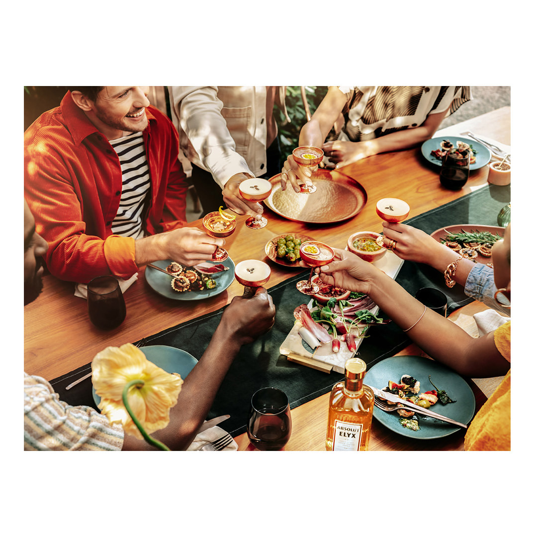 Several people toasting with elegant stemmed cocktail glasses at an outdoor table setting with appetizers and a bottle of liquor.