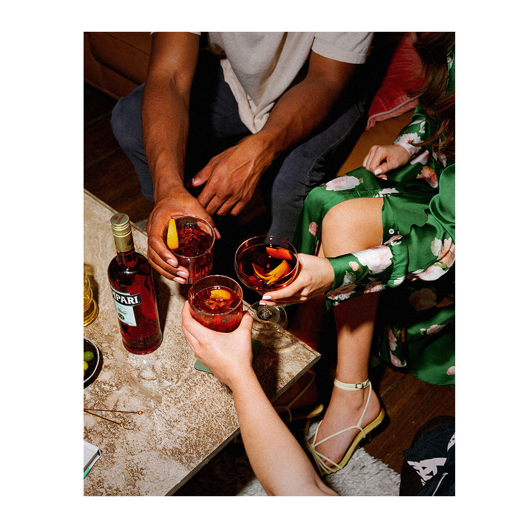 Overhead shot: two people toast with Negroni cocktails beside a Campari bottle. One wears a green floral dress and yellow sandals.