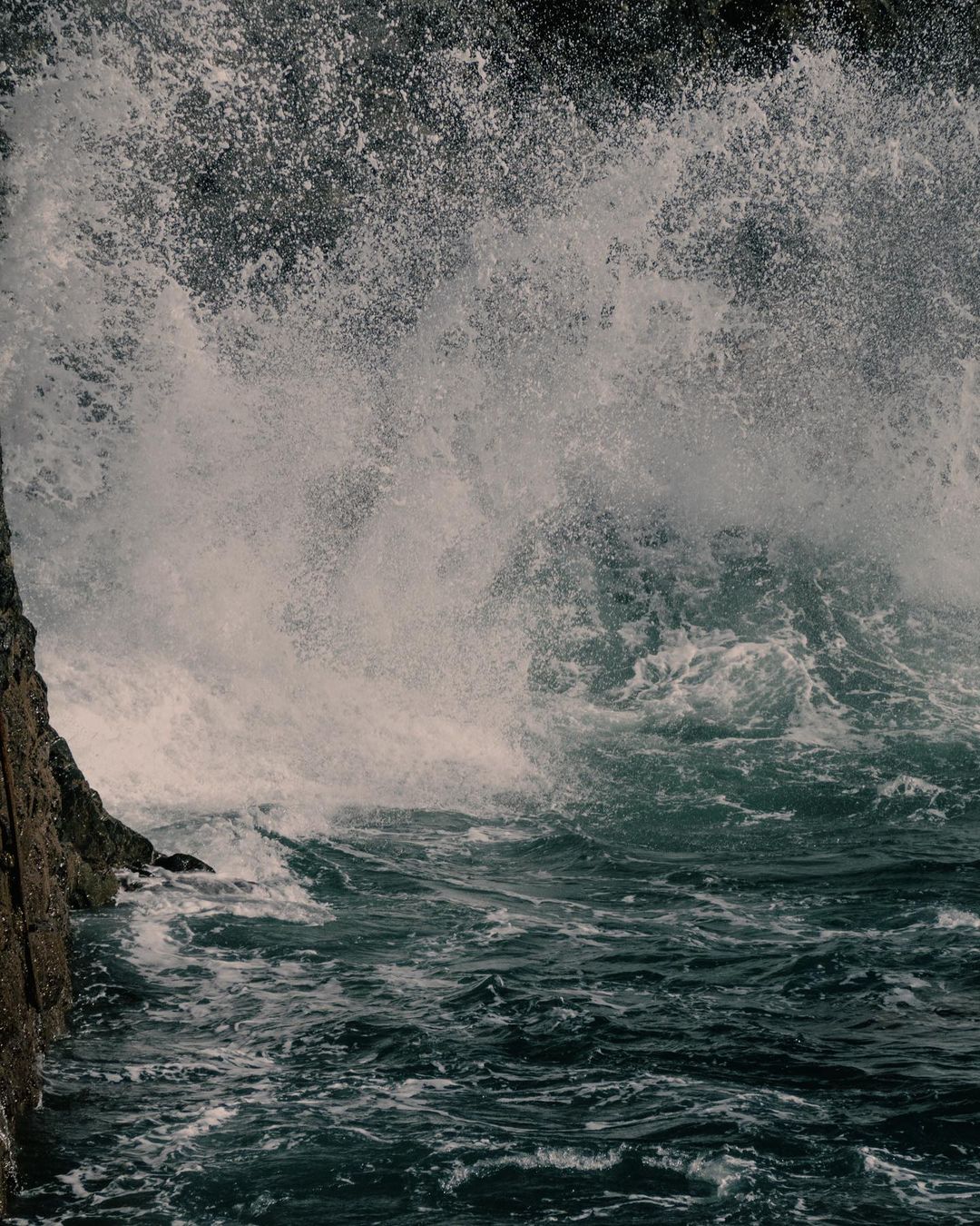 Ocean waves crashing against dark, jagged cliffs, creating a dynamic and textured scene with sea foam and strong contrasts.
