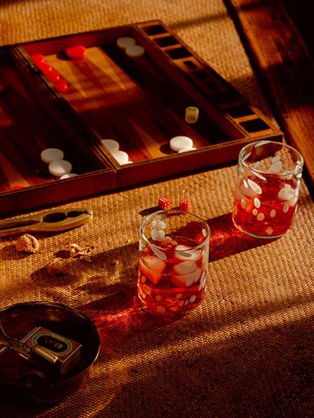 Overhead shot of a backgammon board, dice, glasses with liquid, and objects on a textured brown surface with natural window light.