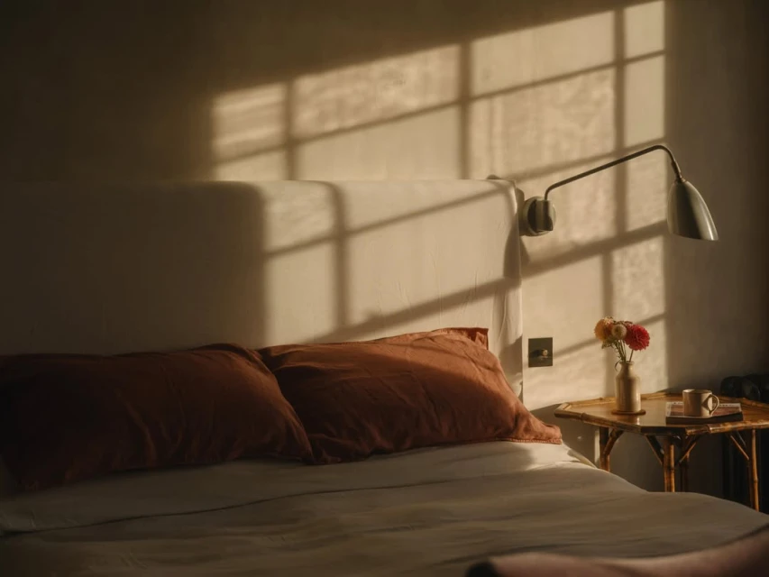 Bedroom interior with bed, side table, flowers, and strong window light creating shadows on the wall in warm tones, cozy room decor.
