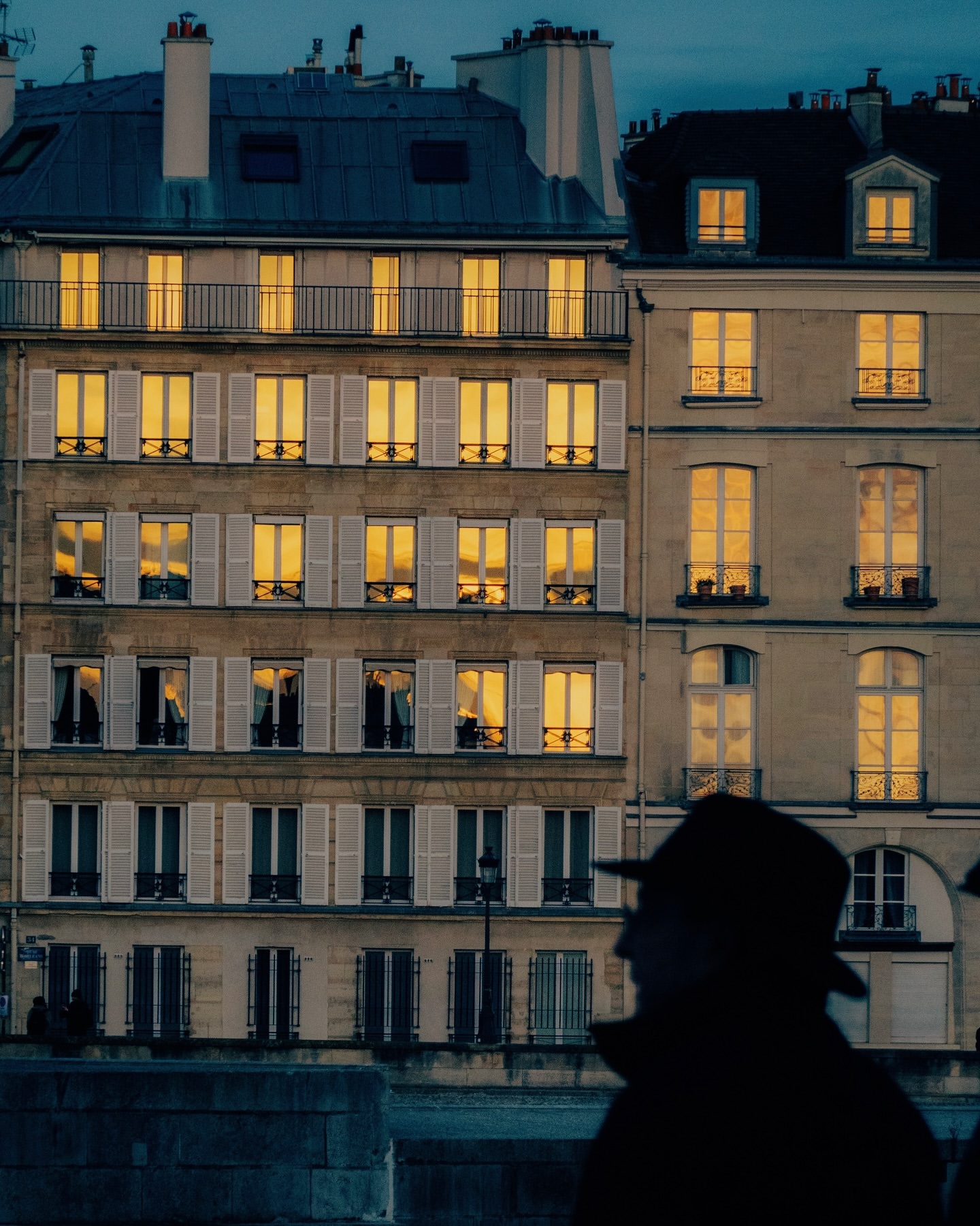 Urban scene with buildings illuminated at dusk, featuring architectural details and silhouette of a person in the foreground. City lights.