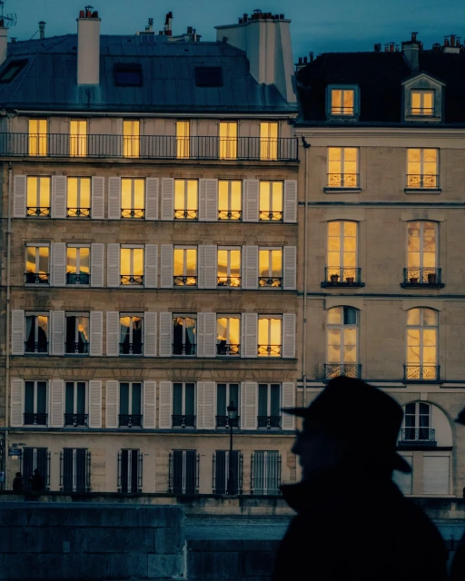 Urban scene with buildings illuminated at dusk, featuring architectural details and silhouette of a person in the foreground. City lights.