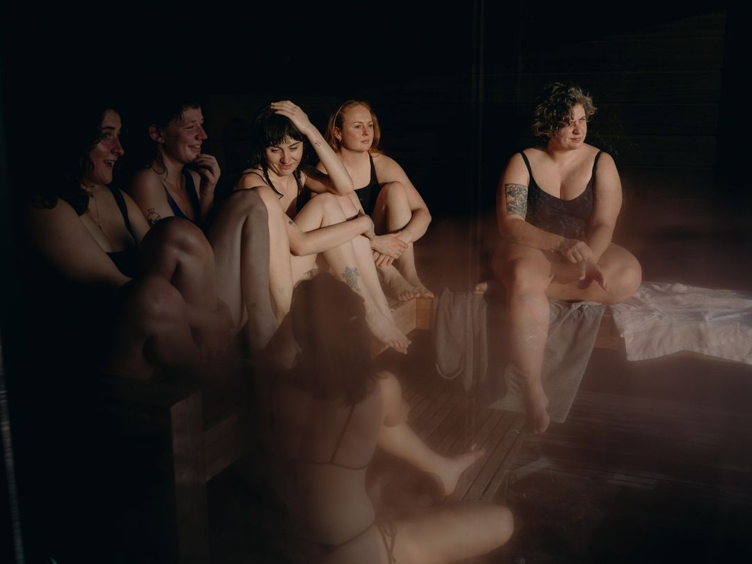 Six women in swimwear sit on wooden benches inside a dimly lit sauna, illuminated by soft natural light, in a casual, intimate gathering.