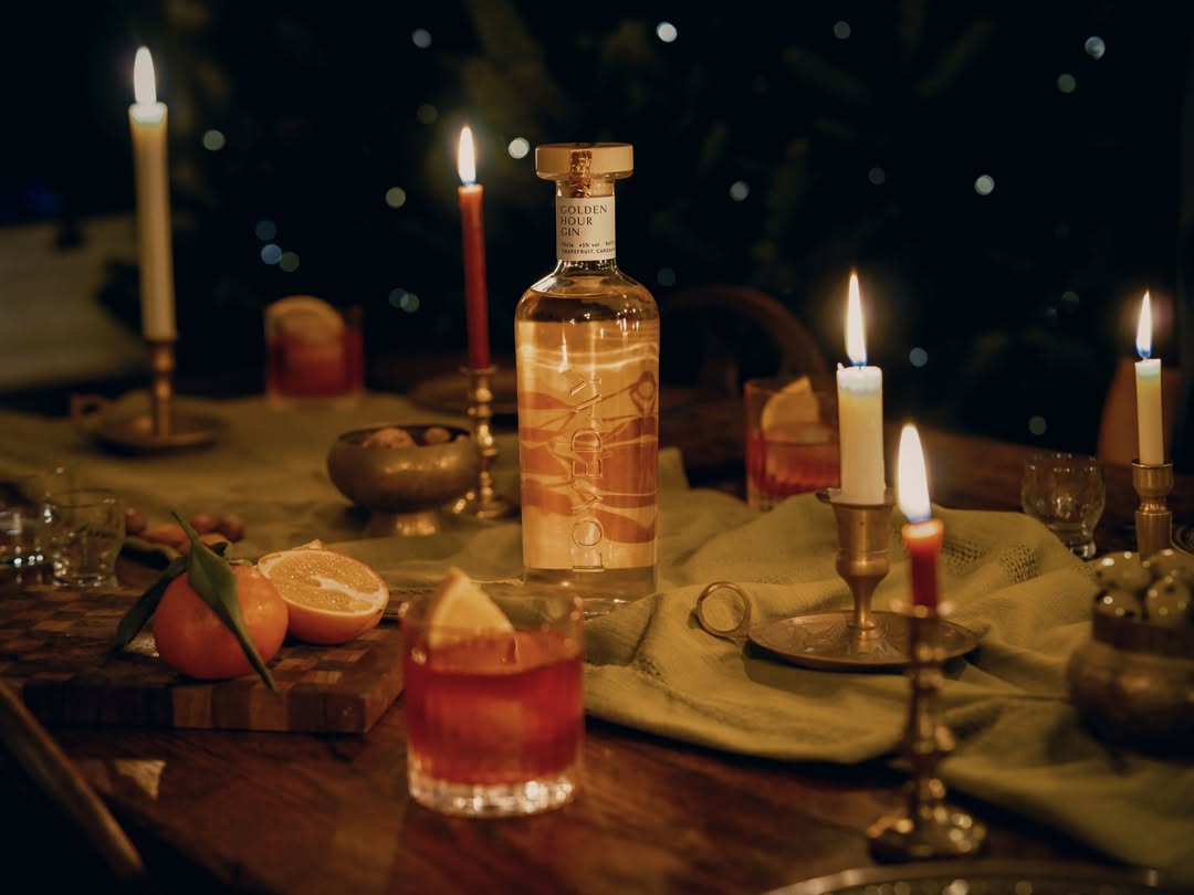 A bottle of gin surrounded by candles, oranges, and cocktail drinks on a wooden table. Warm and inviting ambiance.