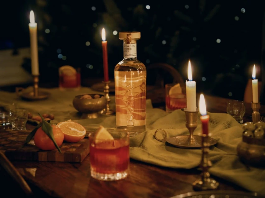 A bottle of gin surrounded by candles, oranges, and cocktail drinks on a wooden table. Warm and inviting ambiance.
