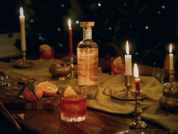 A bottle of gin surrounded by candles, oranges, and cocktail drinks on a wooden table. Warm and inviting ambiance.
