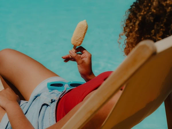 Woman in swimwear lounges by the pool eating an ice cream bar while wearing blue-framed sunglasses on her lap in summer sunlight.