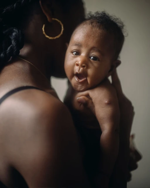 Close-up of an infant being held by an adult; the baby has dark skin, is topless, and has an open mouth; the adult is partially visible.
