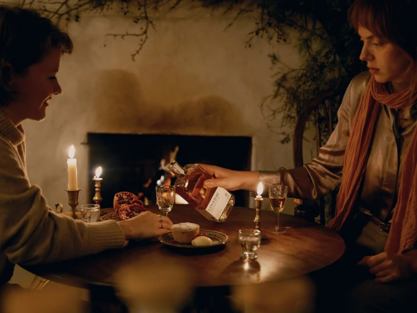Two women at a round table indoors in warm light, pouring alcohol from a bottle into a glass, with food and candles.