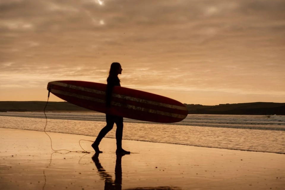 A woman walks on a sandy beach carrying a surfboard, silhouetted against a golden sky at sunset, with ocean waves visible.