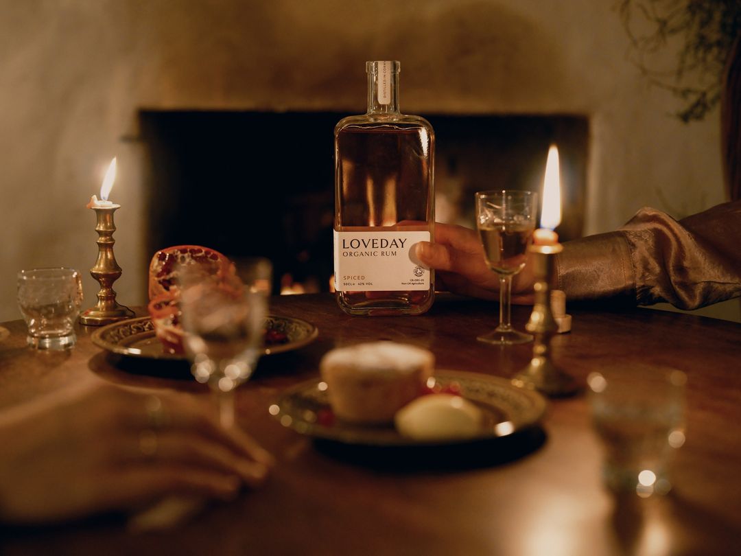 A bottle of LOVEDAY Organic Rum and lit candles are on a wooden table next to food, creating a warm and inviting ambiance.