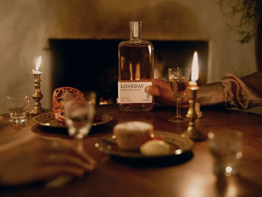 A bottle of LOVEDAY Organic Rum and lit candles are on a wooden table next to food, creating a warm and inviting ambiance.