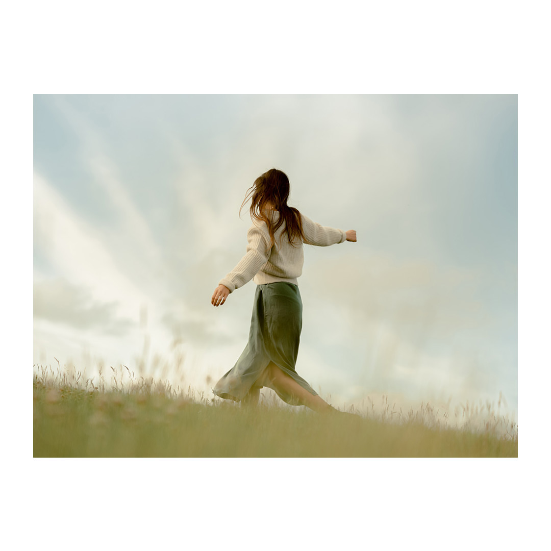 Adult woman with outstretched arms walking on a grassy hillside under a cloudy sky wearing a sweater and skirt in a lifestyle fashion shot.
