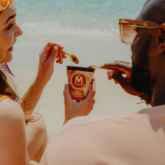 Two people sharing a Magnum Double Sunlover ice cream on a sunny beach, featuring golden spoons and refreshing tropical flavors.