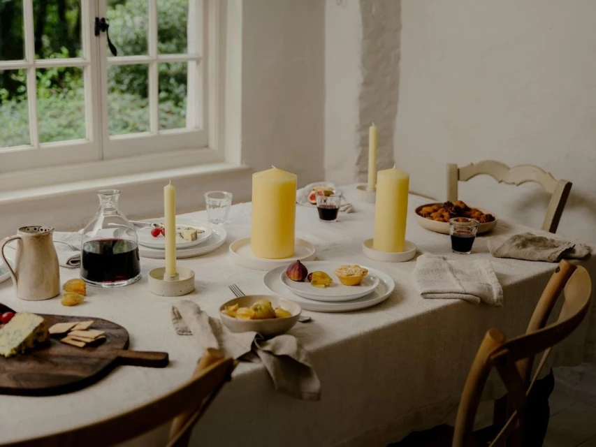 Still life captures a table setting with lit candles, wine, fruit, and snacks, featuring neutral colors and natural light by a window.