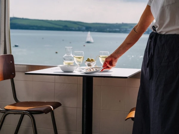 Adult reaches across a white table with a glass of white wine, dish of sardines, and olives in a restaurant with a waterfront view.
