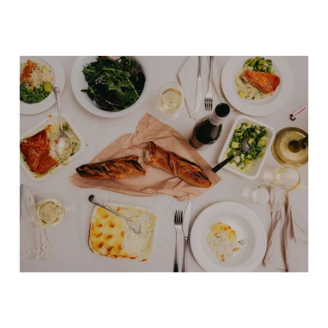 Overhead shot of a dining table covered in plates of food, a bottle of wine, glasses, silverware, and parchment paper with bread.