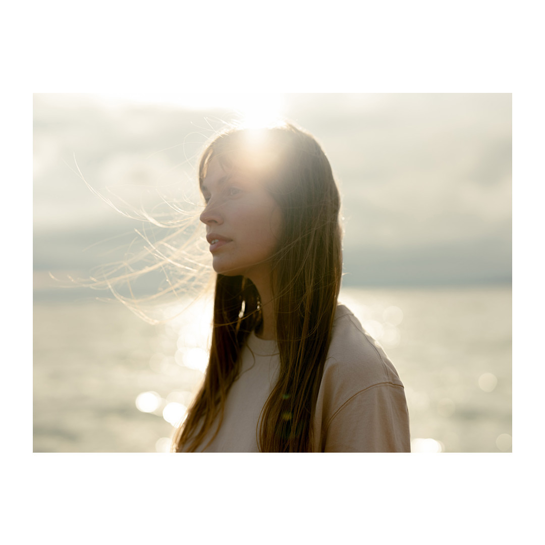 Portrait of a young adult woman with long brown hair standing outdoors near a body of water, the sun shining in the background.