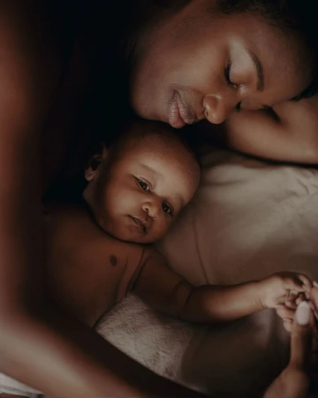 Close-up portrait of a Black mother resting with her infant baby lying on cream bedding in London by Jenna Foxton