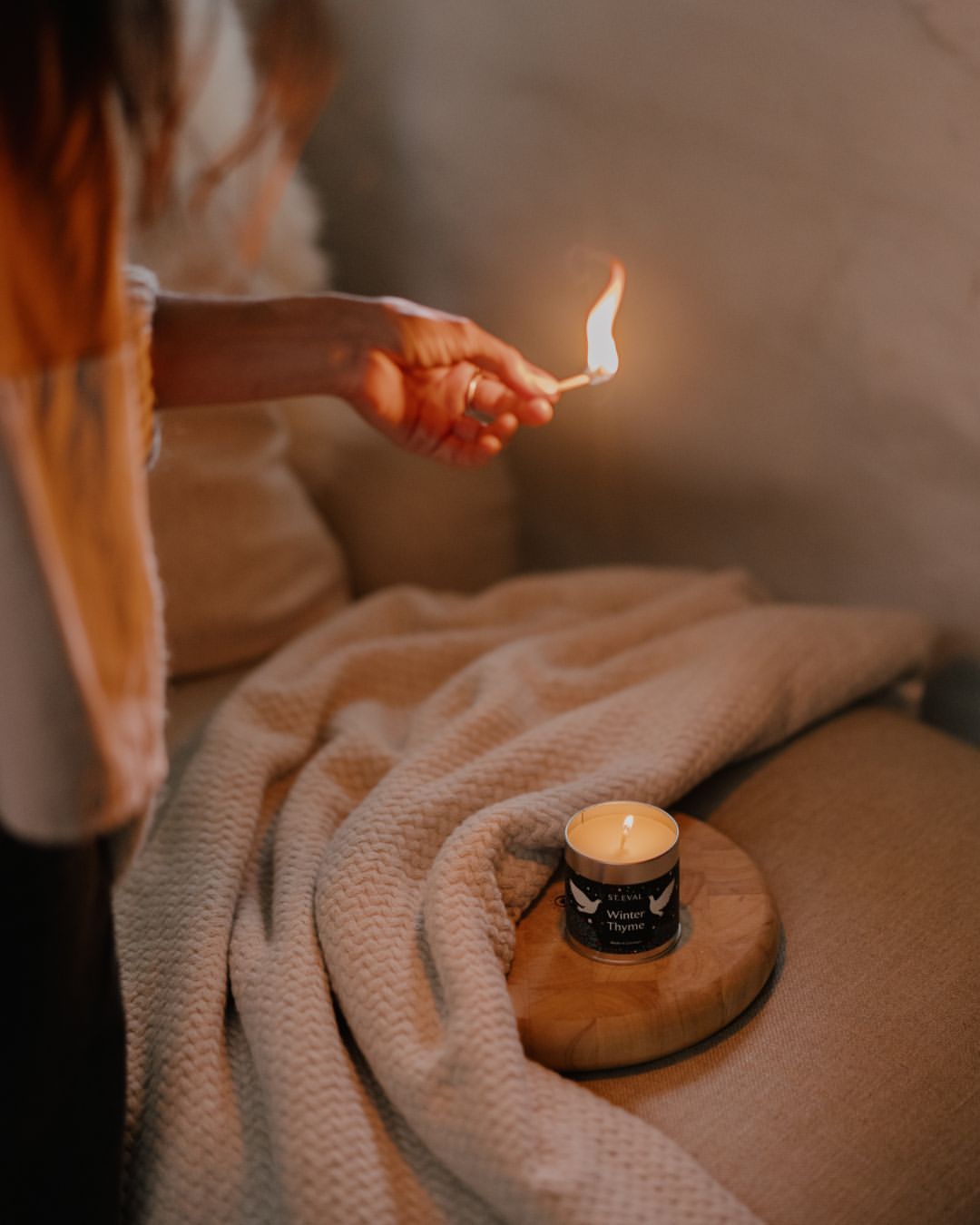 Person lights a candle with a match on a wooden coaster atop a cream-colored textured blanket on a tan sofa in a dimly lit room.