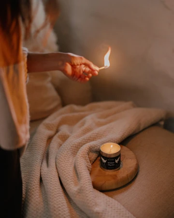 Person lights a candle with a match on a wooden coaster atop a cream-colored textured blanket on a tan sofa in a dimly lit room.