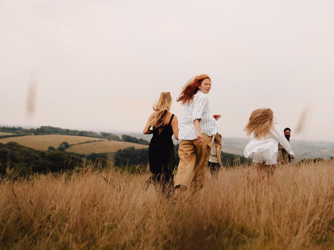 Group of people running through a grassy field with rolling hills in the background, enjoying a day in the countryside, outdoors.