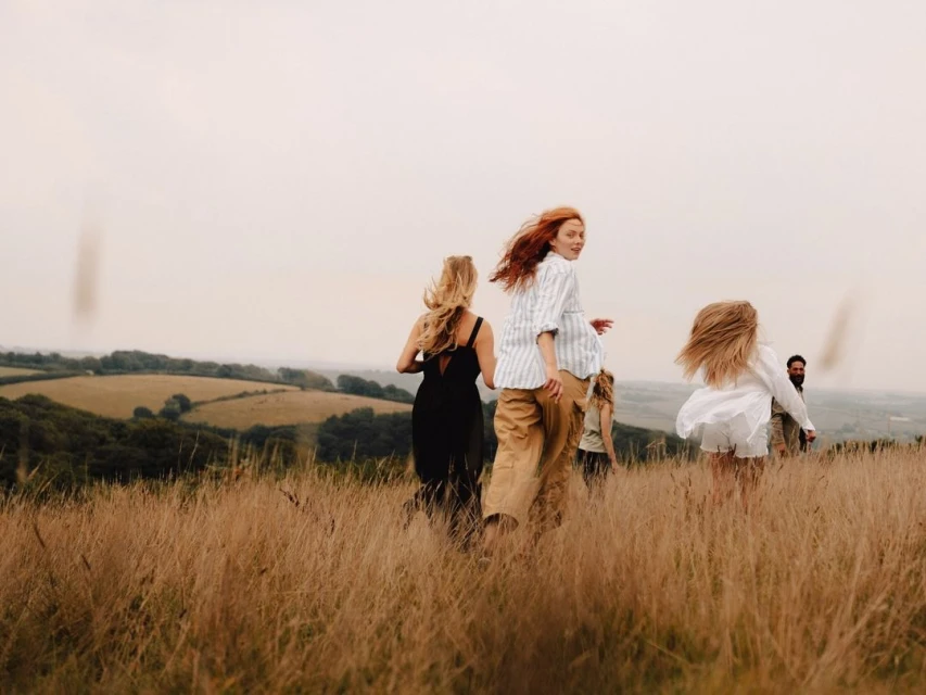 Group of people running through a grassy field with rolling hills in the background, enjoying a day in the countryside, outdoors.