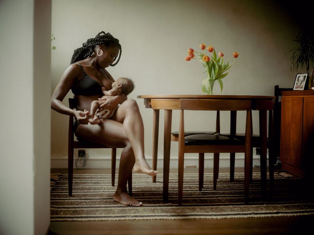 African American woman in black bra breastfeeds a baby indoors near a wood table with tulips, capturing an intimate maternal moment.