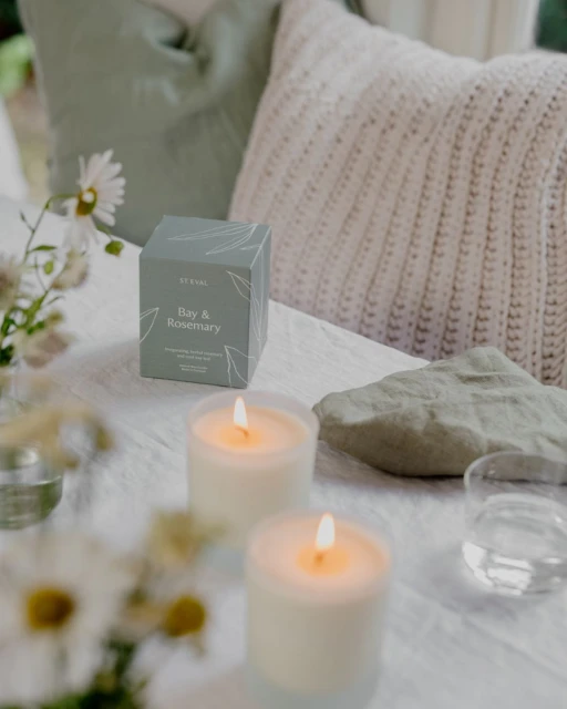 Candles and a 'Bay & Rosemary' gift box sitting on a white cloth with floral arrangements and pillows in the background.