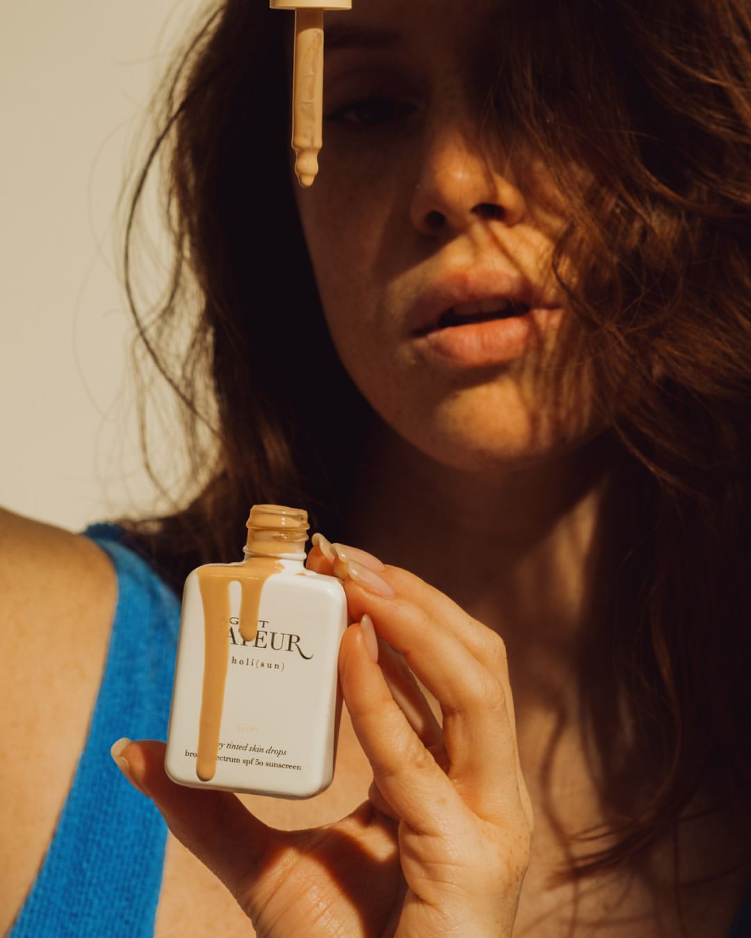 Woman in blue tank top holding a bottle with beige liquid dripping and a dropper, promoting beauty and skincare products.