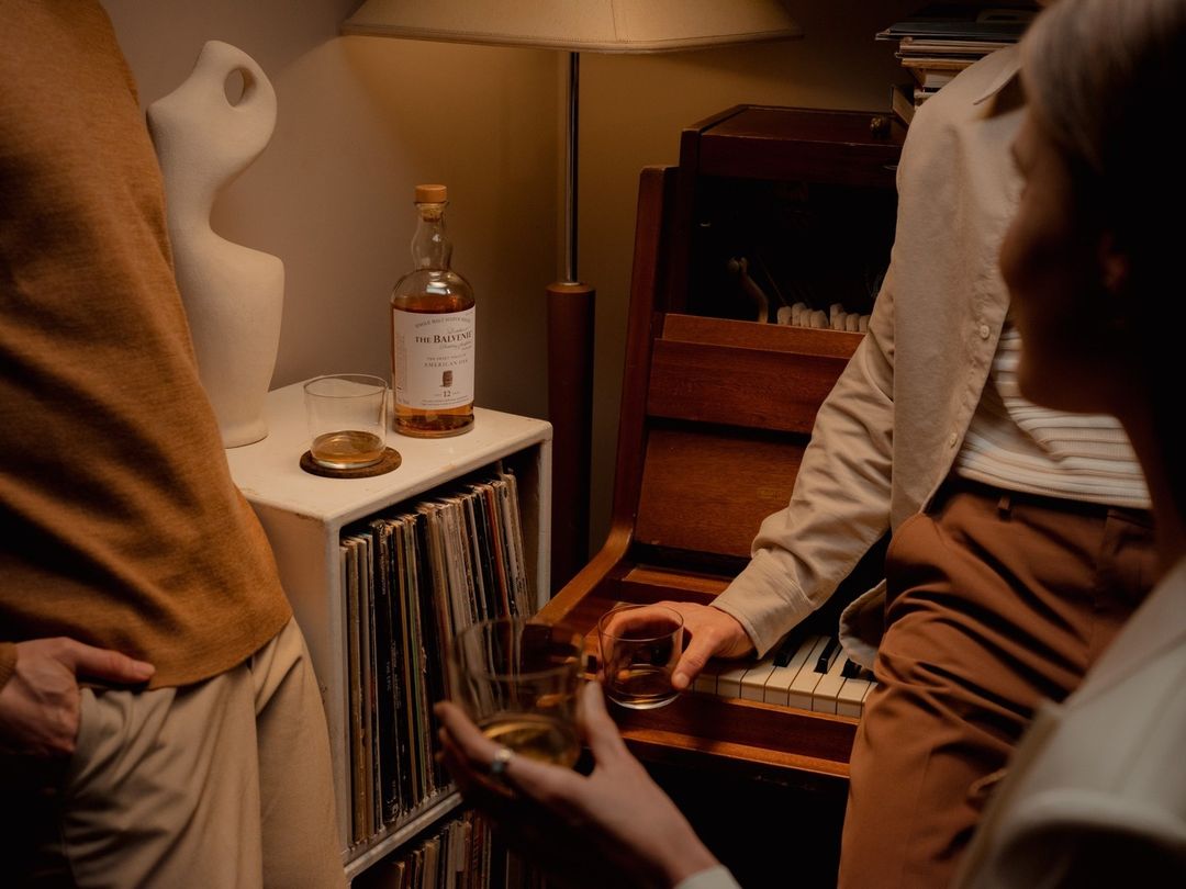 Two adults are indoors, one sitting at a piano with a glass of whiskey, and another standing next to a whiskey bottle on a record cabinet.