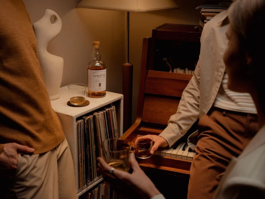 Two adults are indoors, one sitting at a piano with a glass of whiskey, and another standing next to a whiskey bottle on a record cabinet.