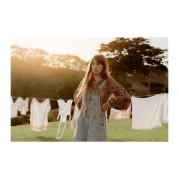 Woman wearing denim overalls standing outdoors in a grassy field with a clothesline behind her.