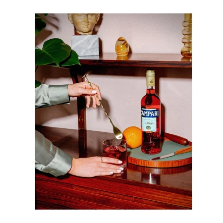 Campari bottle and glass on a wooden sideboard with hands holding a cocktail spoon and orange fruit by Jenna Foxton