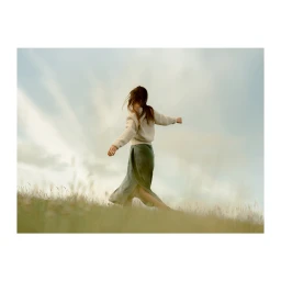 Woman with long brown hair wearing a cream sweater and green skirt spinning in a grassy meadow under a cloudy sky.