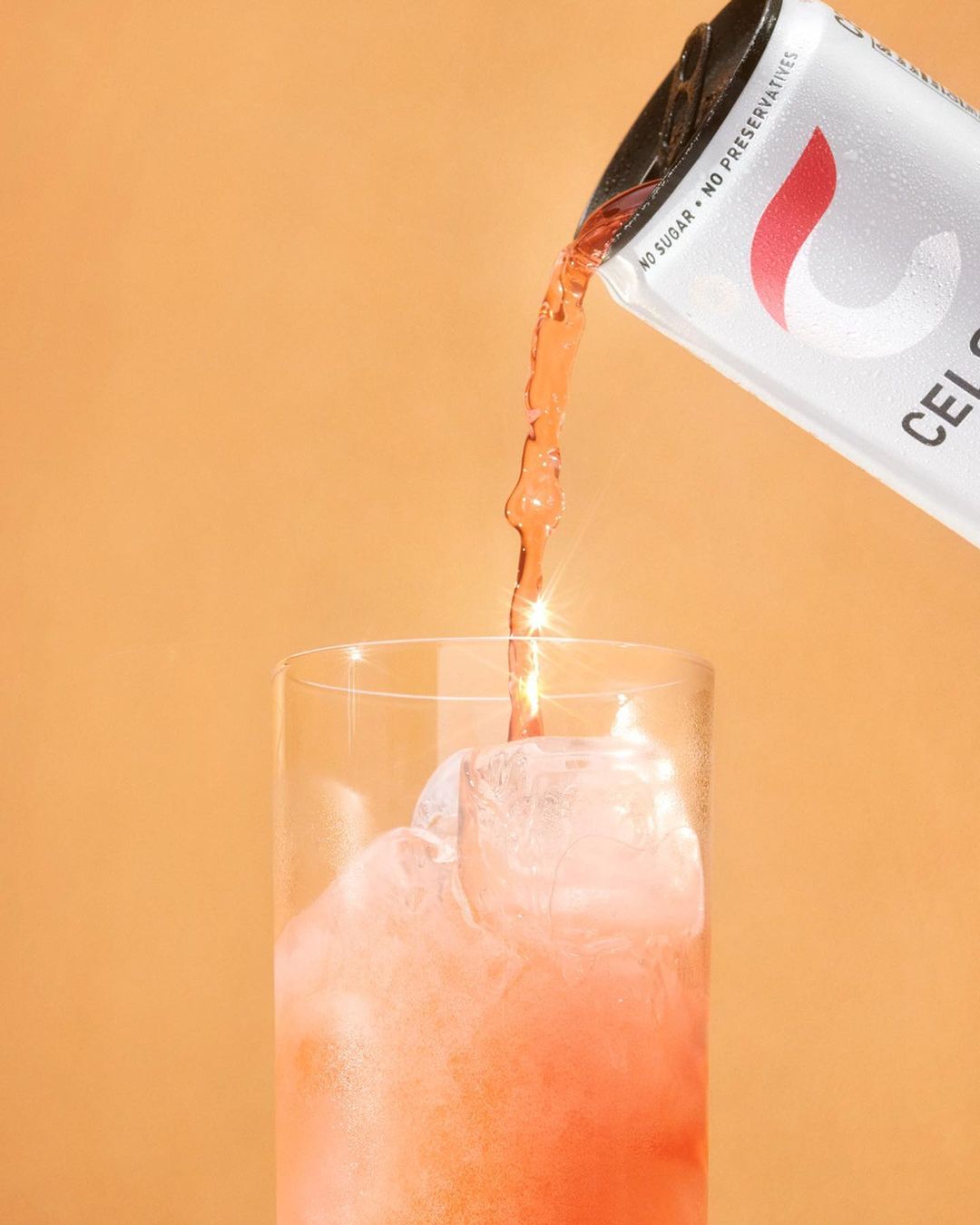 A can of Celsius brand drink is pouring liquid into a tall glass of ice against a peach-colored background, creating a refreshing beverage.