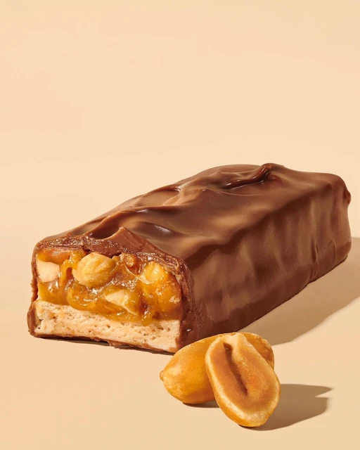 A cross-section of a chocolate bar with peanuts and caramel, presented against a tan background. Two peanuts lie in the foreground.