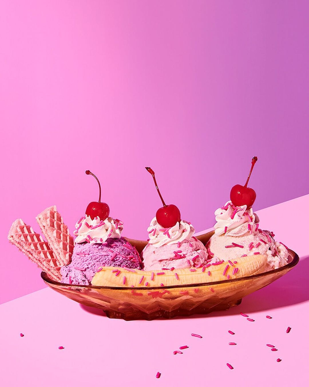 Brightly colored banana split with scoops of ice cream, whipped cream, cherries, and waffle cookies against a pink backdrop. 