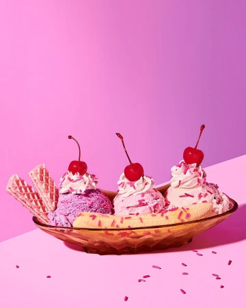 Brightly colored banana split with scoops of ice cream, whipped cream, cherries, and waffle cookies against a pink backdrop. 