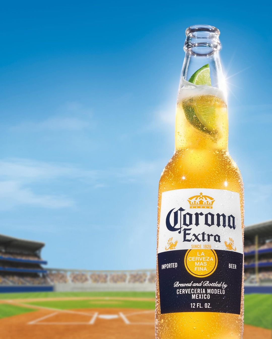 Close-up of a glass bottle of Corona Extra beer with a lime wedge, set against a backdrop of a baseball stadium under a sunny sky.