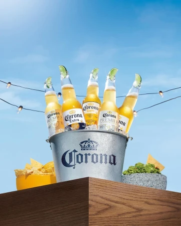 Five Corona beer bottles with lime wedges, chilling in an ice bucket with tortilla chips and guacamole, set against a blue sky backdrop.