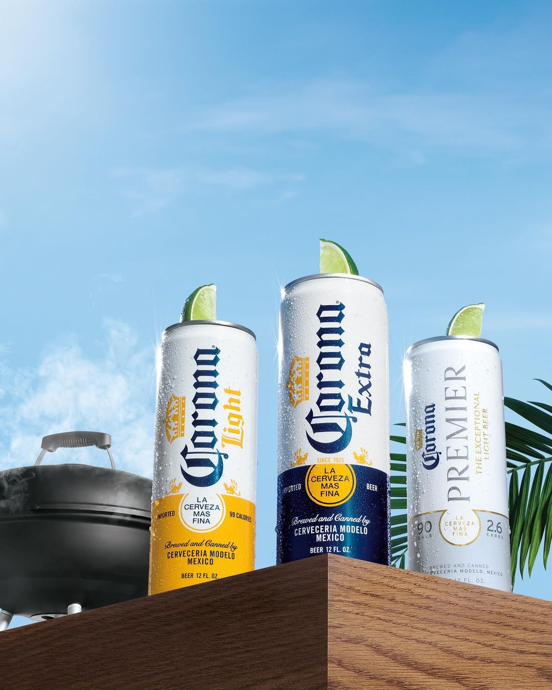 Three cans of Corona beer (Light, Extra, and Premier) on a wooden surface with lime wedges, set against a bright blue sky backdrop.