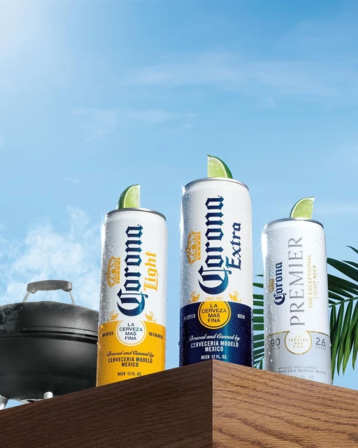 Three cans of Corona beer (Light, Extra, and Premier) on a wooden surface with lime wedges, set against a bright blue sky backdrop.