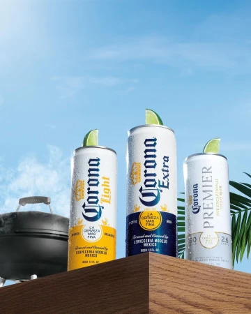 Three cans of Corona beer (Light, Extra, and Premier) on a wooden surface with lime wedges, set against a bright blue sky backdrop.