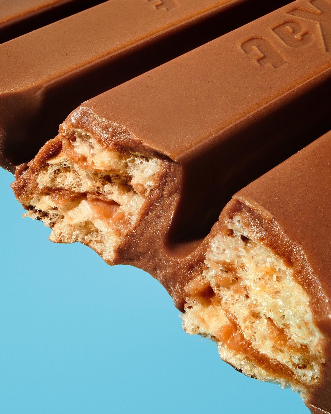 Close-up of a partially eaten Kit Kat chocolate bar against a bright blue background, showing the layers of wafer and chocolate.