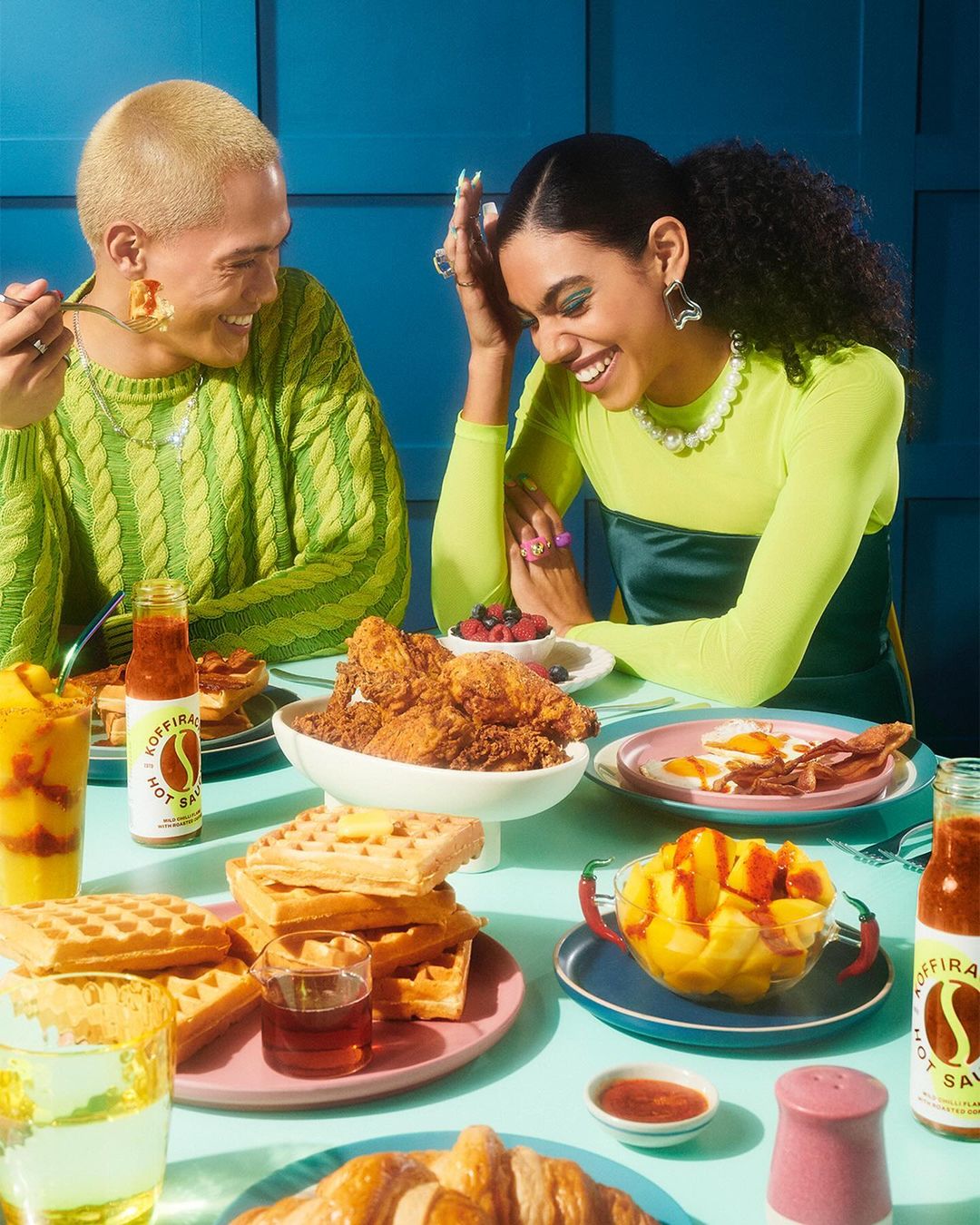 Two people smiling and enjoying a meal of fried chicken, waffles, and fruit, with a bottle of hot sauce on the table.