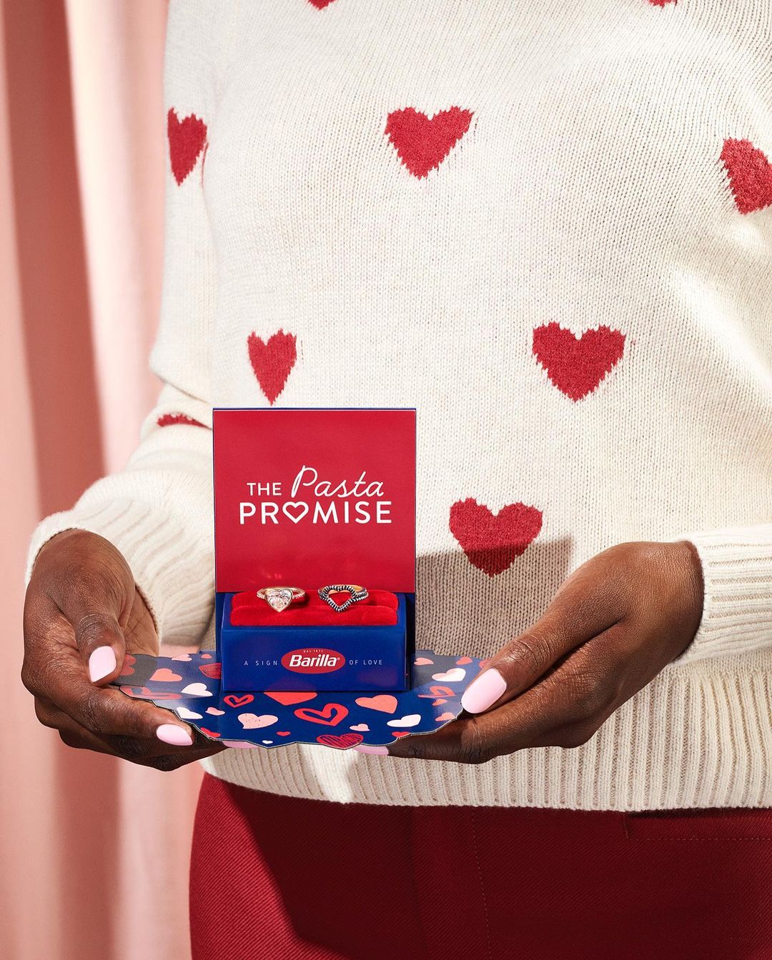 Hands holding a Barilla pasta promise ring box, showcasing two heart-shaped rings against a heart-patterned sweater backdrop.