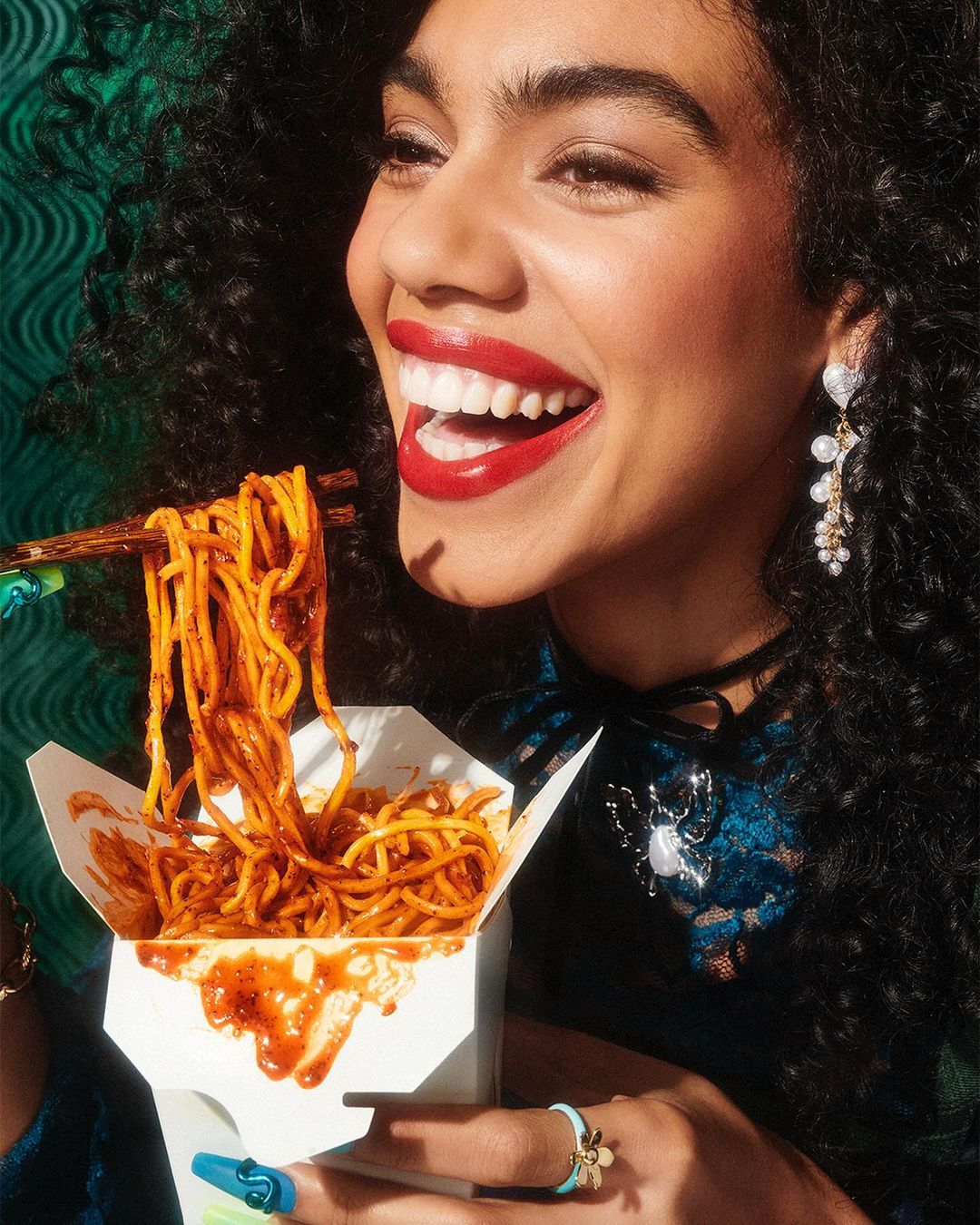 Smiling woman with dark curly hair eating noodles with chopsticks from a take-out container in a brightly lit, close-up studio shot.