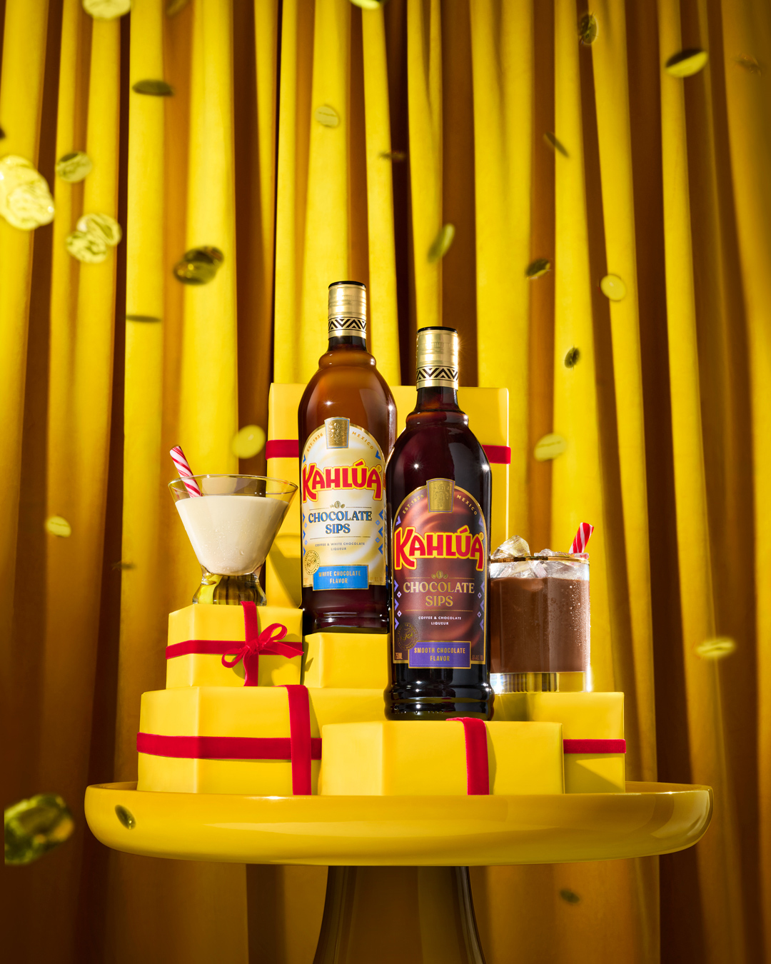 Two Kahlua Chocolate Sips bottles are showcased on yellow gift boxes with red ribbons and a yellow table with a golden curtain backdrop.